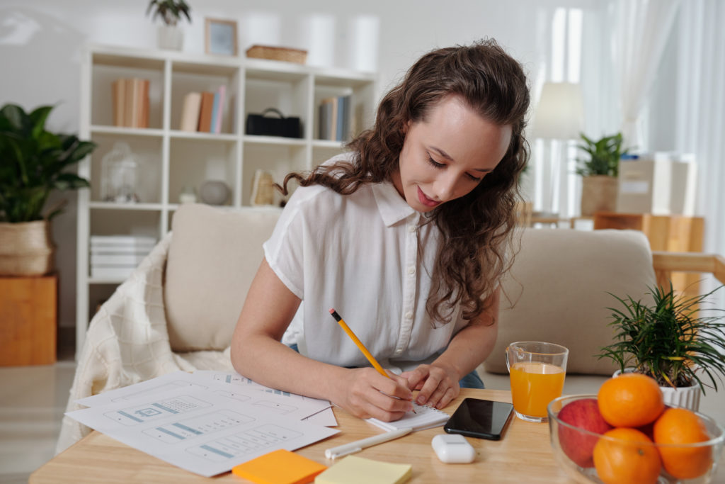 UI designer writing down creative ideas in notepad when working at coffee table at home